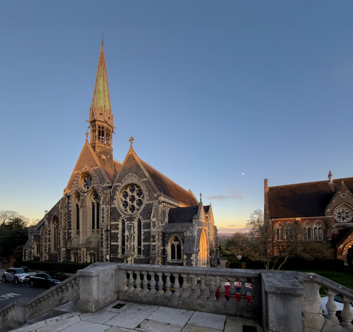 A historical church building made of stone and flint with detailed gothic architectural features, including pointed arches, large stained glass windows, and decorative stone tracery. The church has a tall, slender spire covered in moss and weathered tiles, rising prominently against a clear blue sky during sunset. The surrounding environment includes a paved stone terrace with a traditional balustrade in the foreground, overlooking the church courtyard. Nearby are parked cars and trees with sparse foliage, suggesting late autumn or early winter. The scene is illuminated by soft, natural evening light, accentuating the textures of the stonework and the weathered finish of the church’s historic facade. The setting implies a quiet, peaceful atmosphere typical of a small community or historic area, aligning with the context of independent or private disposal services that Rubbish Removal Harrow offers for waste collection away from public authority schemes.