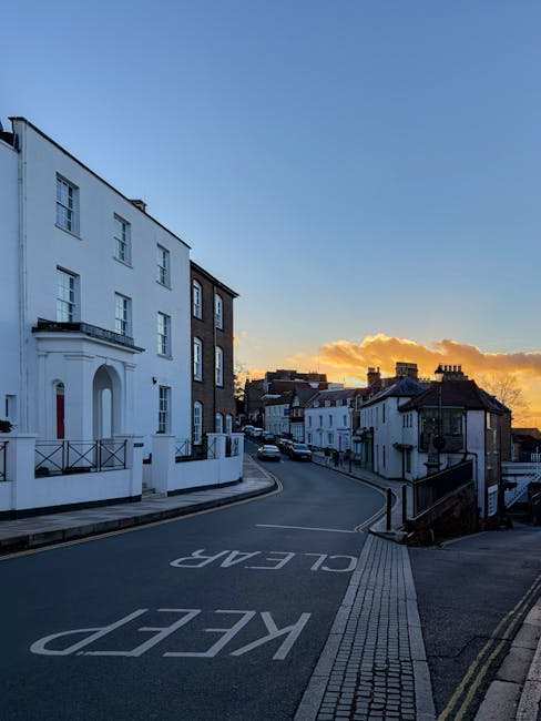 A quiet residential street during sunset, with a large white multi-story building on the left featuring multiple windows and a small balcony with black railings. The road curves gently to the right, marked with large white lettering reading 'KEEP CLEAR' in the foreground. On the right side, a steep pavement with tactile paving runs alongside a sloped, dark-colored metal railing. The street is lined with smaller white and brick buildings, some with tiled roofs, extending into the distance under a sky with wispy clouds illuminated by the setting sun. The scene exudes a calm, urban atmosphere typical of a suburban area, where private waste collection services like those offered by Rubbish Removal Harrow can be conveniently integrated into the regular street layout for efficient rubbish removal and disposal management.