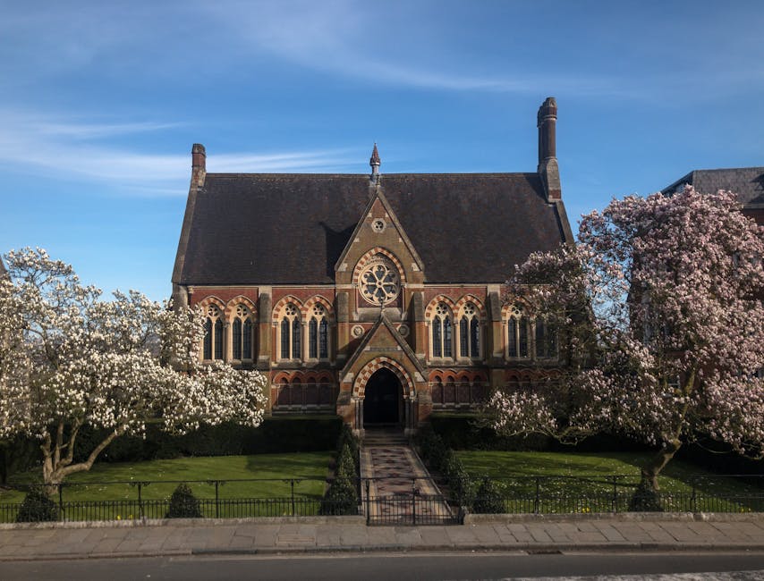 A historical church building made of stone and flint with detailed gothic architectural features, including pointed arches, large stained glass windows, and decorative stone tracery. The church has a tall, slender spire covered in moss and weathered tiles, rising prominently against a clear blue sky during sunset. The surrounding environment includes a paved stone terrace with a traditional balustrade in the foreground, overlooking the church courtyard. Nearby are parked cars and trees with sparse foliage, suggesting late autumn or early winter. The scene is illuminated by soft, natural evening light, accentuating the textures of the stonework and the weathered finish of the church’s historic facade. The setting implies a quiet, peaceful atmosphere typical of a small community or historic area, aligning with the context of independent or private disposal services that Rubbish Removal Harrow offers for waste collection away from public authority schemes.