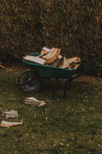 A green plastic wheelbarrow filled with cut logs and pieces of timber, positioned on a grassy outdoor area with a background of dense green shrubbery or hedge. Several smaller wood fragments are scattered on the ground around the wheelbarrow, which has a black metal frame and a single rubber wheel. The logs in the wheelbarrow display a light, natural wood tone with rough, uneven textures, and some show visible grain patterns. The scene is illuminated by natural daylight, creating soft shadows and highlighting the organic textures of the wood and foliage. This image relates to outdoor waste clearing or garden rubbish removal, exemplifying an environment prepared for private disposal or landscaping waste collection, with the wheelbarrow serving as a typical tool for manual rubbish transport. The branding of Rubbish Removal Harrow is subtly present, aligning with the context of professional rubbish services specializing in garden and domestic waste removal.