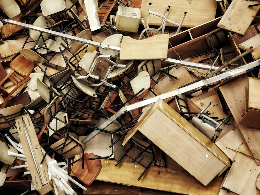 A large pile of assorted discarded furniture and office equipment, including wooden tables, chairs with metal and wooden frames, and a beige filing cabinet, all stacked haphazardly inside a storage or warehouse space. The surfaces of the tables vary from smooth and polished to rough and untreated, with some showing signs of wear or damage. The chairs are of different designs, some with plastic or wooden seats, and bodies made of metal or wood, some stacked or leaning on each other. A metal pole or rail crosses diagonally across the scene, partly resting on the furniture. The environment appears industrial, with a concrete or wooden floor visible beneath the clutter, and the lighting is subdued, casting soft shadows across the items. This scene exemplifies a typical collection of bulk waste items prepared for removal or recycling, highlighting the importance of professional waste management services like those provided by Rubbish Removal Harrow for efficient on-site clearance and private disposal of non-household waste materials.
