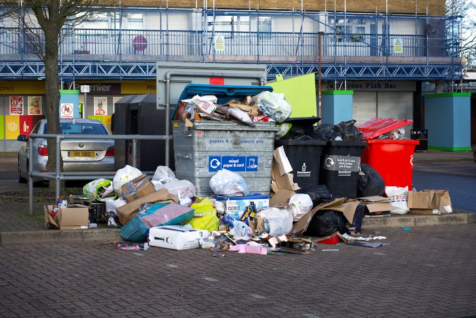 A narrow alleyway filled with accumulated rubbish and waste materials, featuring a large black trash bag and a wheeled bin positioned centrally in the foreground. Behind these, there are numerous cardboard boxes, black plastic bags, and loose debris piled up against a makeshift wall constructed from various discarded items, including wooden pallets and bundled trash. The alley is bordered by tall, weathered buildings on either side, with graffiti visible on the right wall and graffiti on the left side, suggesting an urban environment. Bare tree branches extend over the scene from the background, with a pale, overcast sky casting diffuse lighting over the area. The ground is stained and uneven, reflecting neglect and lack of regular waste clearance. This scene highlights the importance of private rubbish removal services, such as those offered by Rubbish Removal Harrow, for managing waste in urban backstreets and alleyways where street cleaning may not be frequent or sufficient.