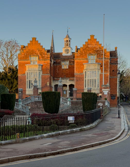 A large historic brick building with a symmetrical facade, featuring prominent stepped gables at each corner and a central clock tower with a weather vane on top. The building's exterior displays red brickwork with contrasting light-colored stone window frames and decorative elements. Tall, narrow bay windows with multiple panes dominate the front, and the structure is surrounded by a small, landscaped garden enclosed by a black wrought iron fence. The garden includes neatly trimmed shrubs and small trees, with a pathway leading up to the building's entrance via a set of stone steps. The building is situated on a curved paved street with a sidewalk and street lamp visible on the right, and a clear blue sky provides natural lighting, accentuating the detailed brickwork and historic architectural features. The scene evokes a sense of preserved heritage, consistent with private or independent property management that may sometimes require rubbish removal services when maintaining or renovating historic sites.