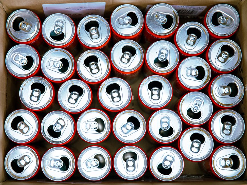 A top-down view of multiple unopened aluminium beverage cans with silver tops and pull tabs, arranged in a dense pattern inside a cardboard box. The cans have a metallic, smooth finish with reflective surfaces, and the pull tabs are uniformly positioned on their top surfaces. The background shows the partially visible brown cardboard sides of the box, which holds the cans tightly together without any additional packaging. The setting appears to be on a flat surface, possibly in a warehouse or collection area for waste collection and recycling purposes. The image captures the cleanliness and uniformity of the cans, emphasizing their material and the way they are stacked for transport or disposal, consistent with a bulk collection of recyclable waste by companies like Rubbish Removal Harrow involved in alternative waste handling and rubbish removal services.
