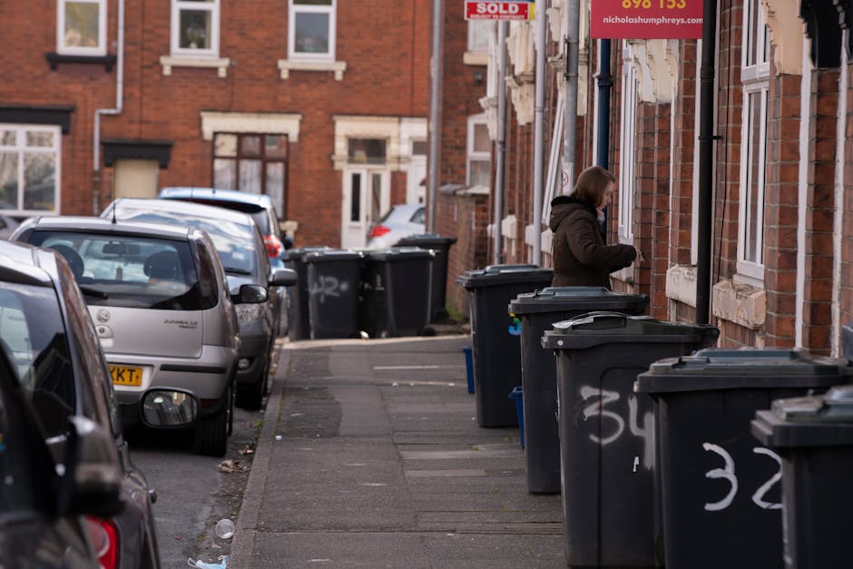 A residential street scene showing a row of black wheelie bins lined up along the pavement outside brick terraced houses. The bins are made of plastic with textured surfaces and are closed, with some lids slightly open. The concrete sidewalk is adjacent to parked cars facing away from the camera, with a few small pieces of litter scattered on the ground. In the background, there are additional bins, more parked vehicles, and brick houses with white-framed windows. One woman, dressed in a dark brown coat, is standing next to a bin, appearing to be engaged in rubbish disposal or collection activity. The environment is lit by natural daylight, suggesting a typical urban or suburban setting suitable for private waste collection services such as those offered by Rubbish Removal Harrow. The scene illustrates a typical rubbish disposal area outside residential properties, aligned with the context of alternative waste handling or on-site clearance.