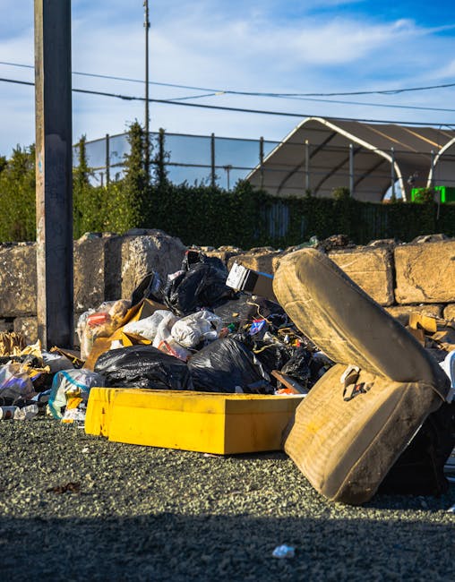 A pile of mixed household waste and rubbish bags, predominantly black plastic, is gathered on a gravel surface adjacent to a stone wall and a metal utility pole. An old, beige car seat with fabric upholstery, showing signs of dirt and wear, is placed in front of the rubbish. The waste includes plastic bags, cardboard, and miscellaneous household debris, with some items spilling onto the ground. In the background, there is a hedge, a partially visible green sports net, and a canopy structure suggesting a sports facility or outdoor court. The scene is illuminated by daylight, with a blue sky and scattered clouds overhead, indicating an outdoor setting in a residential or semi-urban area. The image reflects an example of the type of waste collected through private rubbish removal services, such as those provided by Rubbish Removal Harrow, offering an alternative to council-based refuse collection, especially for bulky items and household debris needing on-site clearance or private disposal.