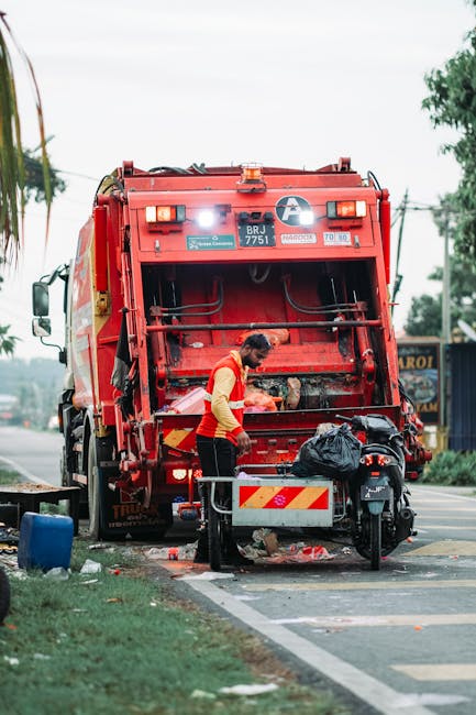 A sanitation worker wearing a yellow high-visibility vest and dark trousers is operating a large red waste collection vehicle, positioned at the edge of a paved street, during daytime. The rear of the vehicle is open, revealing mechanical components and a platform, with the worker leaning forward to handle black plastic trash bags placed on the ground nearby. The scene includes some scattered debris and trash on the pavement, a small table or stand with an orange and white striped safety barrier, and a black motorcycle parked close to the waste truck. In the background, there are trees, utility poles, and a blue building or sign, suggestive of an urban or suburban environment. Natural daylight illuminates the scene, highlighting the textures and colors of the waste collection equipment, the worker’s safety gear, and the surroundings, emphasizing the context of independent collection or alternative waste handling services in a street setting, consistent with rubbish removal activities conducted by companies like Rubbish Removal Harrow.