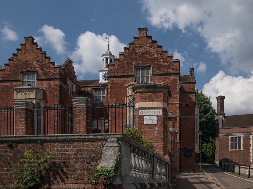 The image depicts a historic red-brick building with Gothic-style architectural features, including stepped gables, pointed arch windows, and decorative stone detailing around the windows and roofline. The building is set behind a black wrought iron fence, supported by brick pillars, with a concrete and brick boundary wall in the foreground. The structure's finish is a mixture of weathered and clean brickwork, with some areas showing aged mortar and others appearing freshly restored. A small white sign on one of the brick pillars indicates the location as 'Church Hill,' and another smaller sign beneath it displays an illegible inscription. The building's roofline features a small white clock tower topped with a weather vane, and the sky above is partly cloudy, with patches of blue visible through the clouds. Surrounding greenery adds contrast to the brickwork, and a narrow lane runs along the side of the building. The scene is lit by natural daylight, creating a clear and detailed view of the historic structure, which may be associated with a church or institutional purpose. The context of rubbish removal services relates to the area as an example of private property where waste clearance could be an alternative to traditional council collection, highlighting opportunities for on-site clearance or independent disposal solutions by companies like Rubbish Removal Harrow.