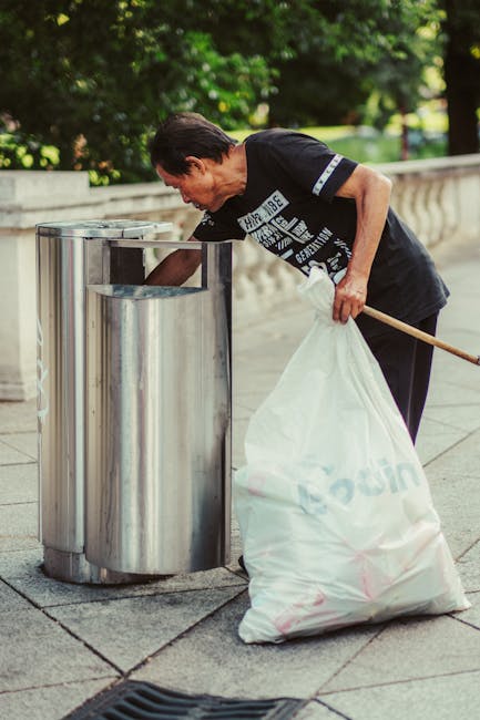 A man with dark hair, wearing a black T-shirt with white graphic text, is seen bending over and placing a large white plastic bag filled with rubbish into a stainless steel public waste bin. The bin has multiple compartments and is situated on a paved sidewalk. Behind him, there are lush green trees and well-maintained bushes, suggesting a park or urban outdoor area. The scene is illuminated with natural daylight, highlighting the outdoor environment and the textures of the metal bin and the man's clothing. The image reflects an act of private waste disposal or rubbish collection in a public space, aligning with waste management or rubbish removal services such as those provided by Rubbish Removal Harrow, showing a clear example of on-site rubbish handling.