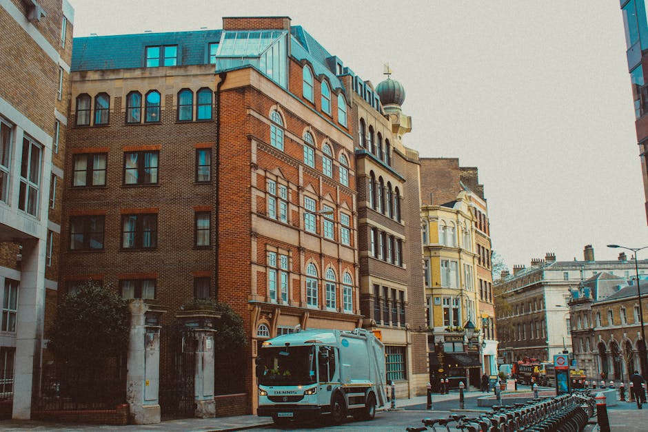 In the foreground, a light blue waste collection truck with a clean, smooth metallic surface and tinted windows is positioned on a city street. The truck is parked near a row of black adjacent bins, which are lined up along the curb, indicating on-site waste collection activity. Behind the truck, a series of multi-story buildings with distinct architectural styles dominate the scene. The central building features a reddish-brown brick facade with arched and rectangular windows framed in white, topped with a teal-colored mansard roof. To the right, a pale yellow building with ornate decorative elements and large sash windows adds visual interest, while further along, buildings of brick and stone exhibit traditional European city architecture. The street is paved with asphalt, and elements like lampposts, a traffic sign, and bicycles are visible, contributing to an urban environment typical of Harrow. The sky appears overcast, casting diffused light over the scene, with minimal shadows, reflecting a quiet moment of private waste management within the cityscape, subtly aligning with rubbish removal services and alternative waste handling options provided by companies like Rubbish Removal Harrow.