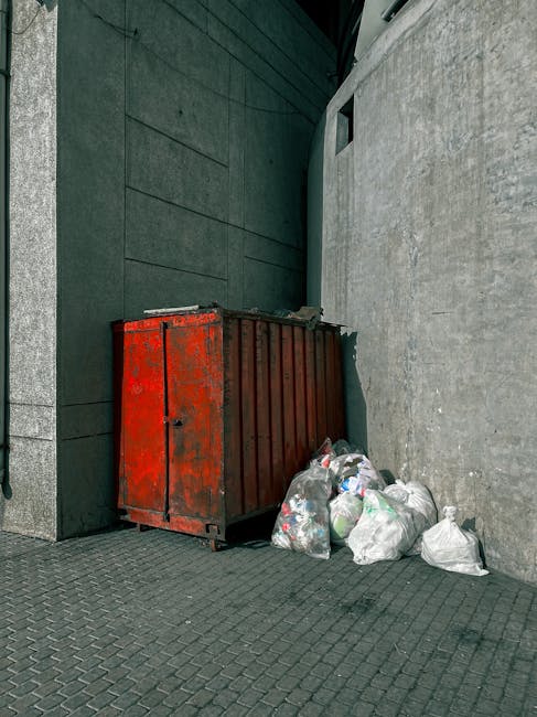 A man with dark hair, wearing a black T-shirt with white graphic text, is seen bending over and placing a large white plastic bag filled with rubbish into a stainless steel public waste bin. The bin has multiple compartments and is situated on a paved sidewalk. Behind him, there are lush green trees and well-maintained bushes, suggesting a park or urban outdoor area. The scene is illuminated with natural daylight, highlighting the outdoor environment and the textures of the metal bin and the man's clothing. The image reflects an act of private waste disposal or rubbish collection in a public space, aligning with waste management or rubbish removal services such as those provided by Rubbish Removal Harrow, showing a clear example of on-site rubbish handling.