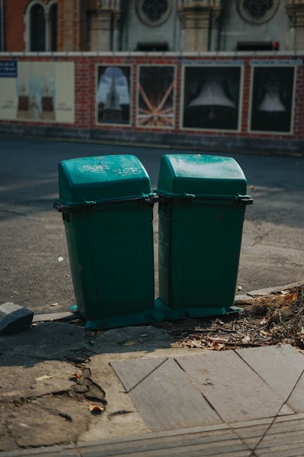 Two green plastic rubbish bins with domed, slightly curved lids are positioned side by side on a paved sidewalk at the edge of a street. The bins are made of durable, textured plastic and are marked with white text and symbols indicating waste disposal. They stand on a small, black plastic base, with some dried leaves and dirt scattered around their bases. Behind the bins, the street surface is asphalt, with a partially visible curb and a building façade featuring decorative brickwork and large, arched windows, suggesting an urban environment. The scene is captured in natural daylight, casting soft shadows on the ground, emphasizing the practical aspect of waste collection services such as those offered by Rubbish Removal Harrow, which may include private waste management or alternative rubbish removal options. The overall setting reflects routine urban waste disposal, with the bins ready for rubbish collection or disposal, aligned with local waste management practices. The image focuses on the functional aspect of street-side waste bins, illustrating their location in a typical urban environment, consistent with professional rubbish removal and waste handling services.