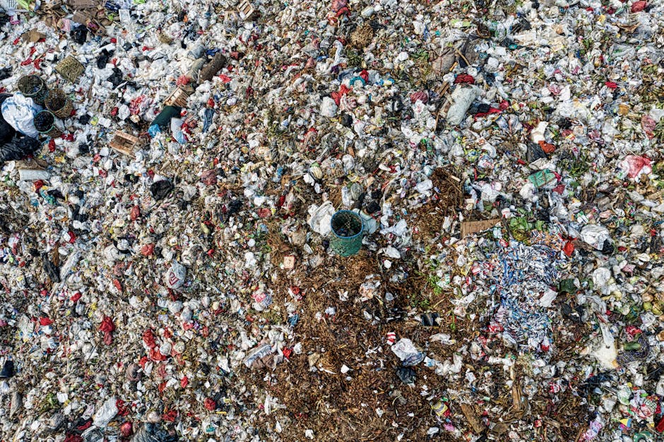 An expansive view of a large landfill site with a thick layer of mixed household and commercial waste, including plastic bags, paper, cardboard, and miscellaneous debris. The waste appears compacted and piled high across the area, with varying textures from crumpled, torn, and flattened materials. Scattered throughout are visible items such as plastic bottles, food packaging, and small fragments of recyclable materials, some partially buried in the heap. A small green plastic bucket is situated on the surface, surrounded by more rubbish, indicating active or recent material placement. The environment is outdoors with natural daylight illuminating the scene, highlighting the chaos of the waste accumulation. This image exemplifies the type of waste that might require professional rubbish removal services, such as those offered by Rubbish Removal Harrow, as an alternative or supplement to local authority waste disposal options and promotes awareness of waste management at large-scale sites.