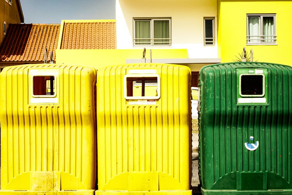 Two green plastic rubbish bins with domed, slightly curved lids are positioned side by side on a paved sidewalk at the edge of a street. The bins are made of durable, textured plastic and are marked with white text and symbols indicating waste disposal. They stand on a small, black plastic base, with some dried leaves and dirt scattered around their bases. Behind the bins, the street surface is asphalt, with a partially visible curb and a building façade featuring decorative brickwork and large, arched windows, suggesting an urban environment. The scene is captured in natural daylight, casting soft shadows on the ground, emphasizing the practical aspect of waste collection services such as those offered by Rubbish Removal Harrow, which may include private waste management or alternative rubbish removal options. The overall setting reflects routine urban waste disposal, with the bins ready for rubbish collection or disposal, aligned with local waste management practices. The image focuses on the functional aspect of street-side waste bins, illustrating their location in a typical urban environment, consistent with professional rubbish removal and waste handling services.